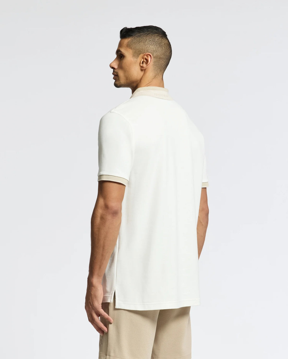 A man with short hair, slightly turned away, showcases the Psycho Bunny MENS NEW CASTLE PIQUE POLO (B6K487F200) in cream. Made of 100% Pima Cotton, it drapes elegantly with his beige shorts and features an embroidered Bunny patch on the plain white backdrop.