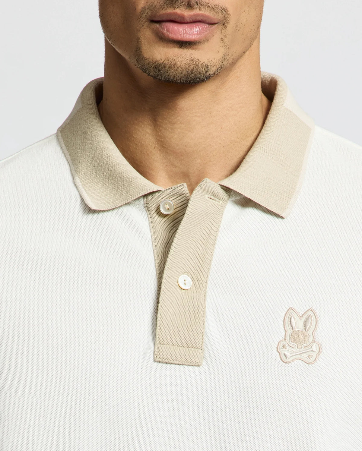 A man sports the MENS NEW CASTLE PIQUE POLO in beige from Psycho Bunny, crafted from 100% Pima Cotton for supreme comfort. It features an embroidered chest Bunny patch on the collar and a two-button placket, set on a plain background.