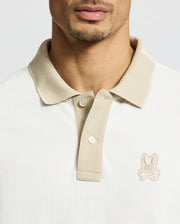 A man sports the MENS NEW CASTLE PIQUE POLO in beige from Psycho Bunny, crafted from 100% Pima Cotton for supreme comfort. It features an embroidered chest Bunny patch on the collar and a two-button placket, set on a plain background.