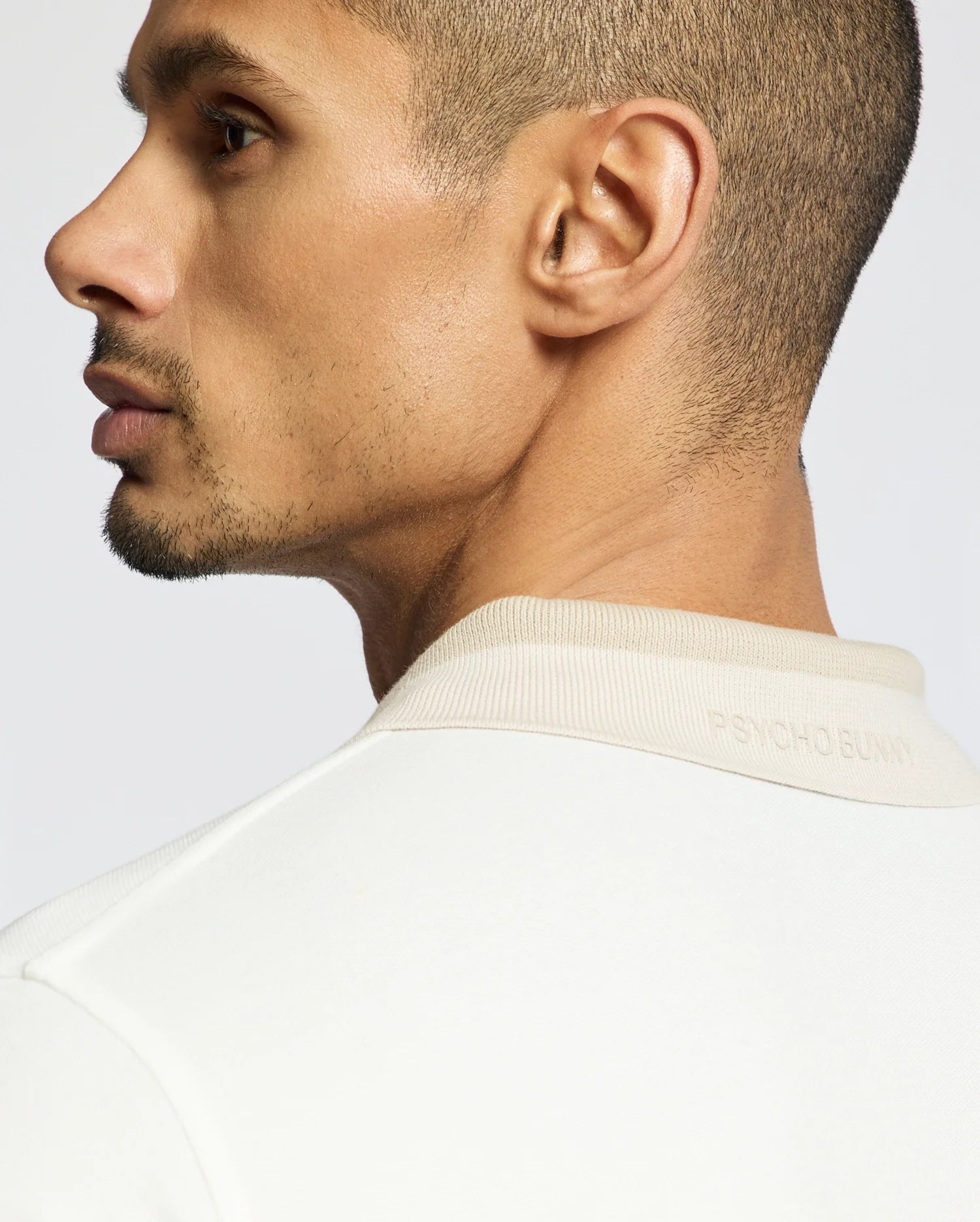 A close-up captures a serious-looking short-haired individual in a cream-colored Psycho Bunny Men's New Castle Pique Polo - B6K487F200, made from 100% Pima Cotton. The collar text subtly enhances the elegant design against a light gray backdrop.