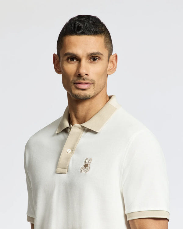 A man wears the Psycho Bunny Men's New Castle Pique Polo (B6K487F200) in 100% Pima Cotton, with a beige collar and bunny logo on his chest. With short dark hair, he gazes at the camera against a plain light background.
