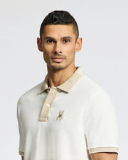A man wears the Psycho Bunny Men's New Castle Pique Polo (B6K487F200) in 100% Pima Cotton, with a beige collar and bunny logo on his chest. With short dark hair, he gazes at the camera against a plain light background.