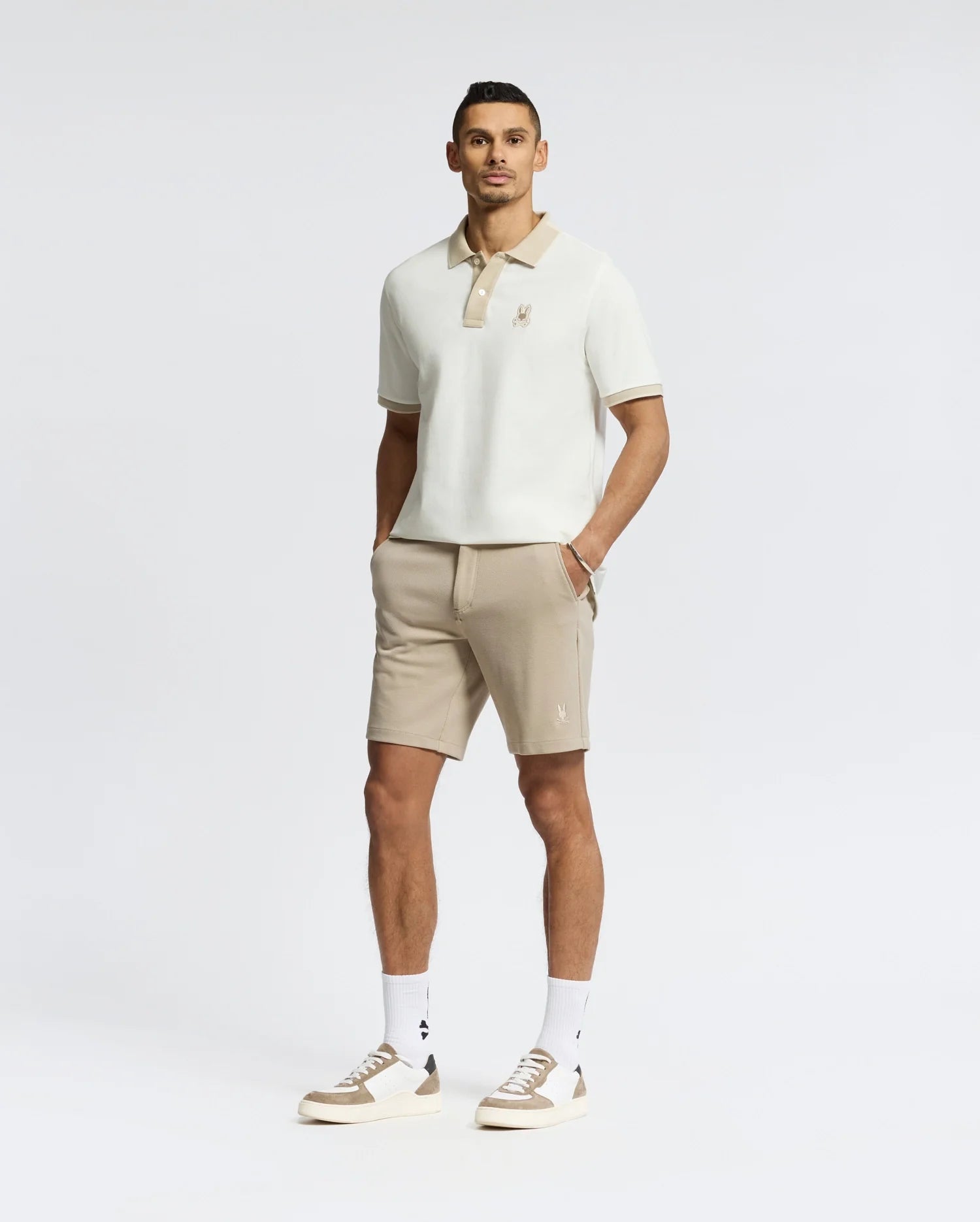 A man stands against a white background, hands in pockets, wearing Psycho Bunny's New Castle Polo (B6K487F200) with beige trim, beige shorts, and white sneakers. The polo features a subtle chest Bunny patch and is made from 100% Pima cotton.