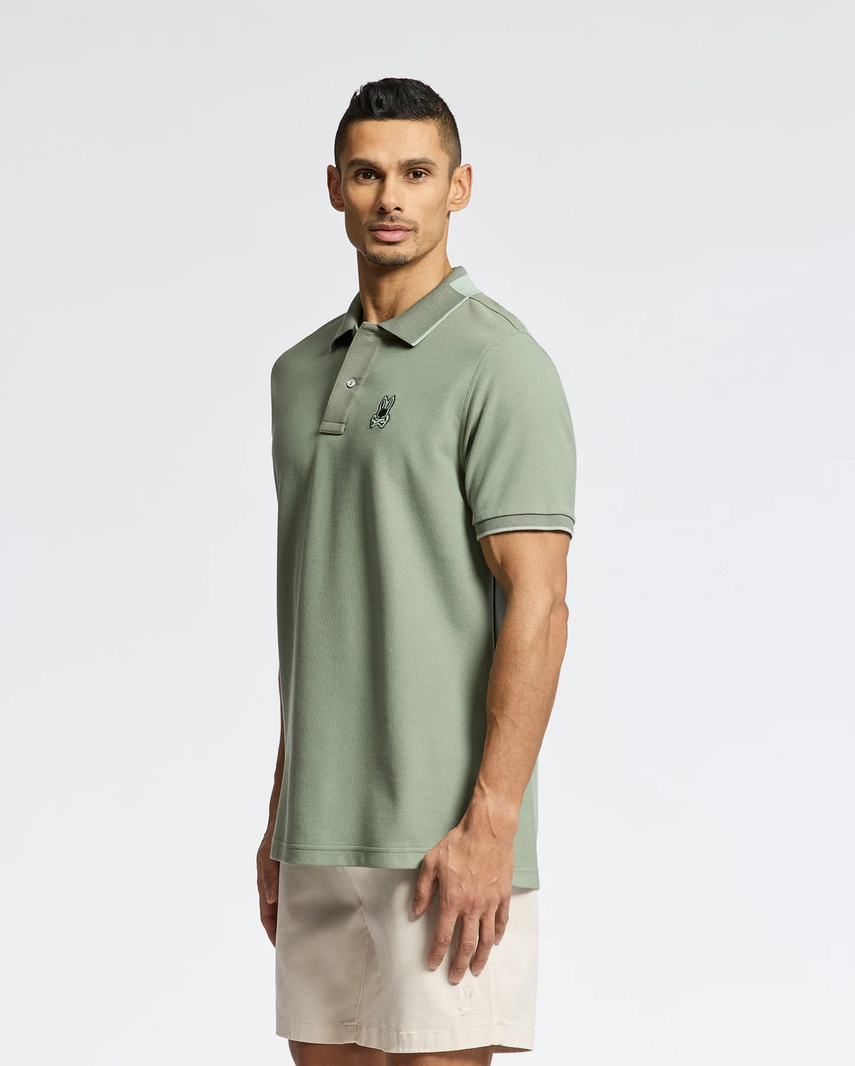 A man with short dark hair and a neutral expression stands against a plain background, wearing the light green, slim fit Psycho Bunny MENS NEW CASTLE PIQUE POLO crafted from luxurious Pima cotton with subtle contrast detailing and a small chest emblem. He's paired it with beige shorts.