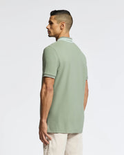 A person with short dark hair is wearing a Psycho Bunny MENS NEW CASTLE PIQUE POLO (B6K487F200) in light green with contrast detailing, paired with beige pants. They stand sideways against a plain white background.