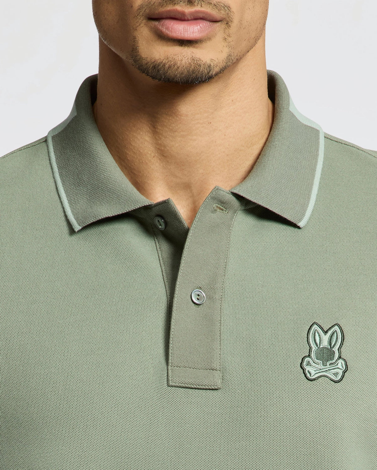 A man wears a Psycho Bunny MENS NEW CASTLE PIQUE POLO (B6K487F200), a slim fit green shirt made from soft Pima cotton, featuring a small cartoon bunny emblem. Its collar and buttons offer classic appeal with stylish contrast detailing on the edges.