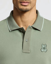 A man wears a Psycho Bunny MENS NEW CASTLE PIQUE POLO (B6K487F200), a slim fit green shirt made from soft Pima cotton, featuring a small cartoon bunny emblem. Its collar and buttons offer classic appeal with stylish contrast detailing on the edges.