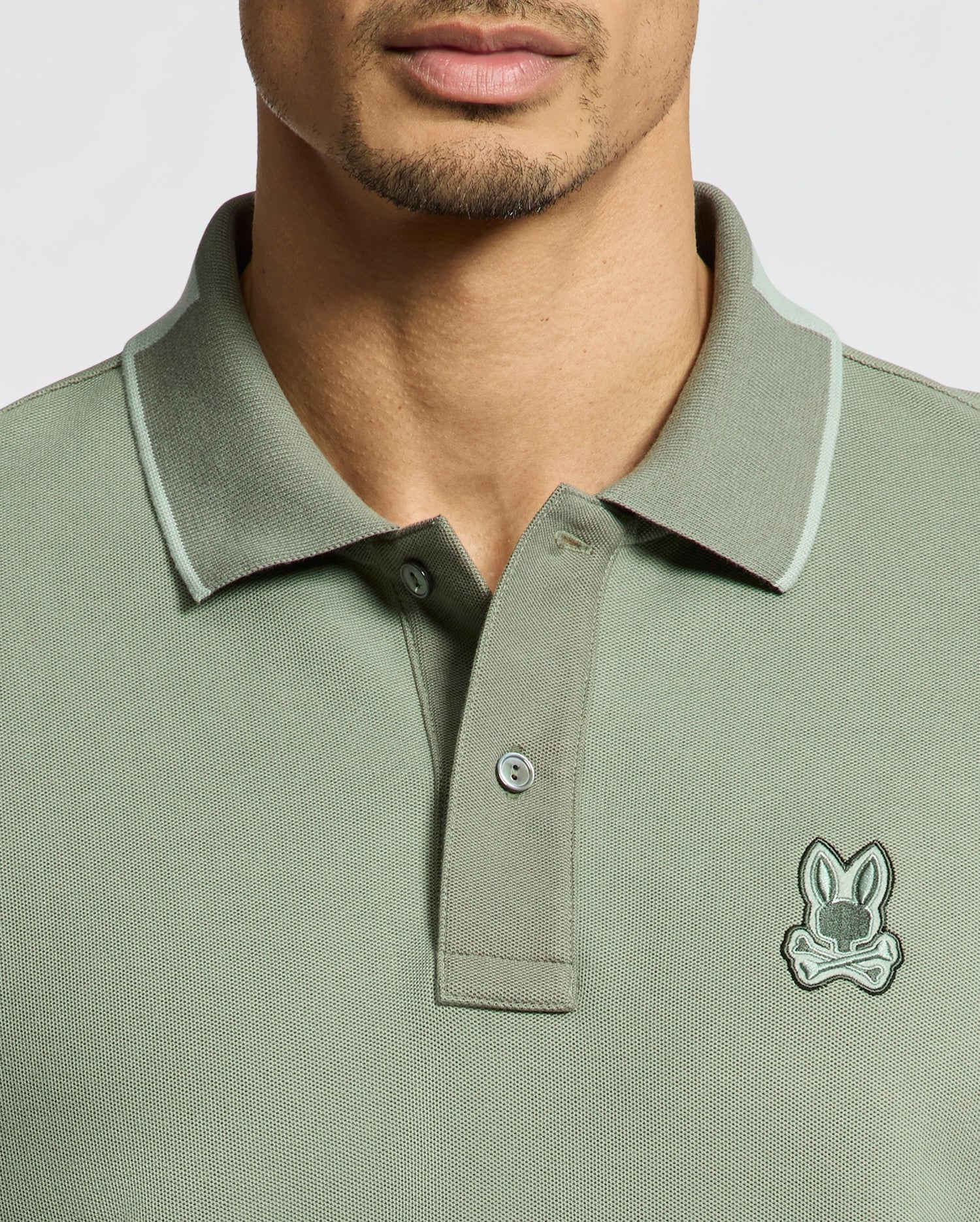 A man wears a Psycho Bunny MENS NEW CASTLE PIQUE POLO (B6K487F200), a slim fit green shirt made from soft Pima cotton, featuring a small cartoon bunny emblem. Its collar and buttons offer classic appeal with stylish contrast detailing on the edges.