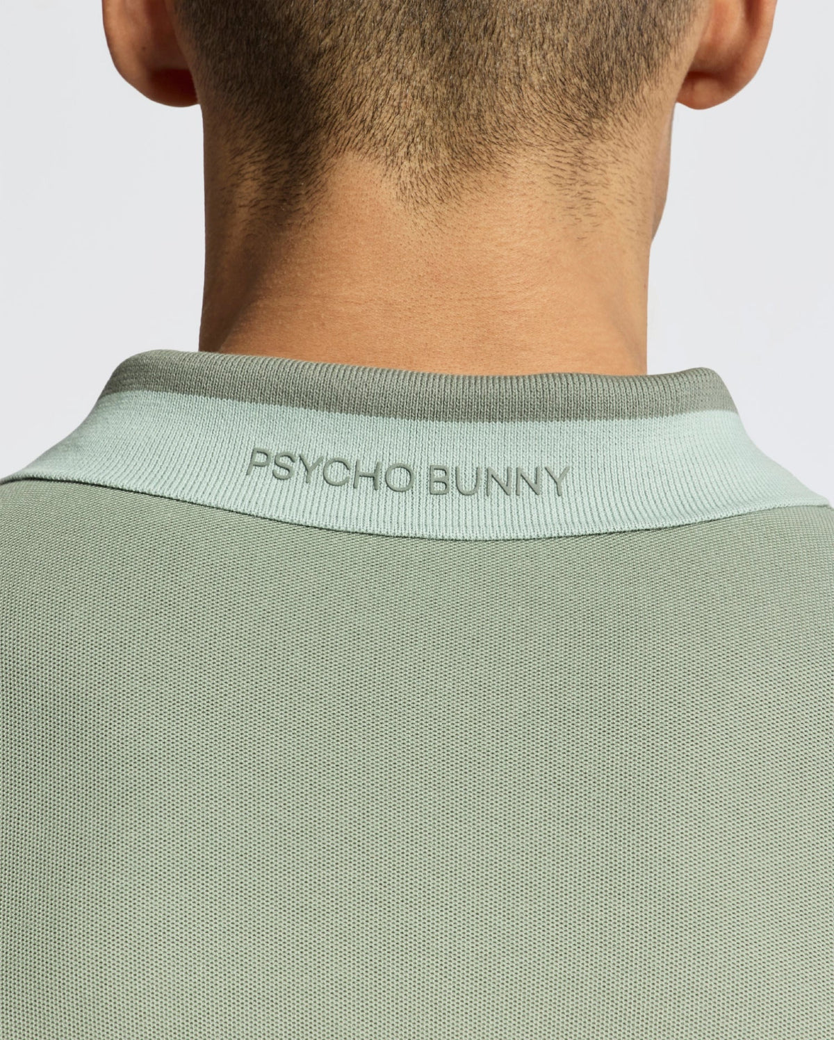 Back view of person in the MENS NEW CASTLE PIQUE POLO - B6K487F200 by Psycho Bunny, a slim fit green Pima cotton shirt with a light green collar and "Psycho Bunny" text embroidered on the back. The plain backdrop enhances its chic appeal with subtle contrast detailing.