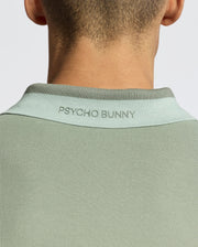 Back view of person in the MENS NEW CASTLE PIQUE POLO - B6K487F200 by Psycho Bunny, a slim fit green Pima cotton shirt with a light green collar and "Psycho Bunny" text embroidered on the back. The plain backdrop enhances its chic appeal with subtle contrast detailing.