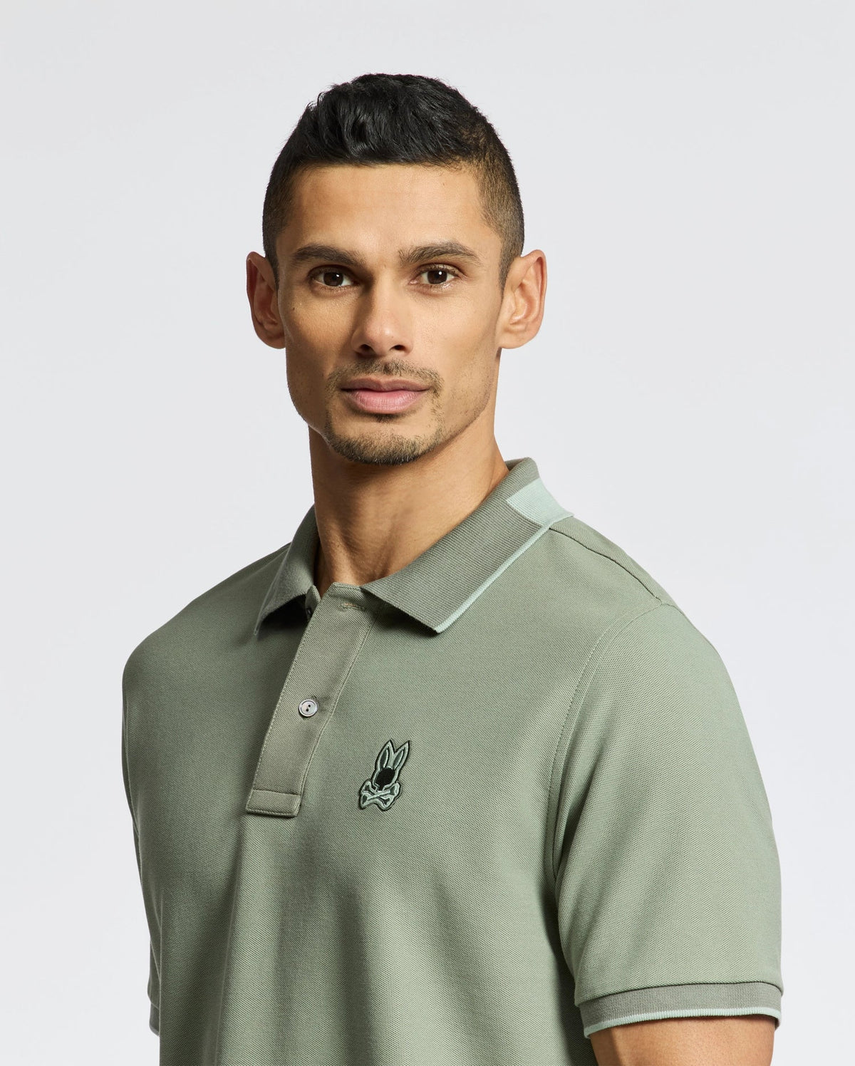 A person with short dark hair wears a muted green slim fit MENS NEW CASTLE PIQUE POLO by Psycho Bunny, crafted from Pima cotton with a small embroidered logo on the chest. The background is plain white.