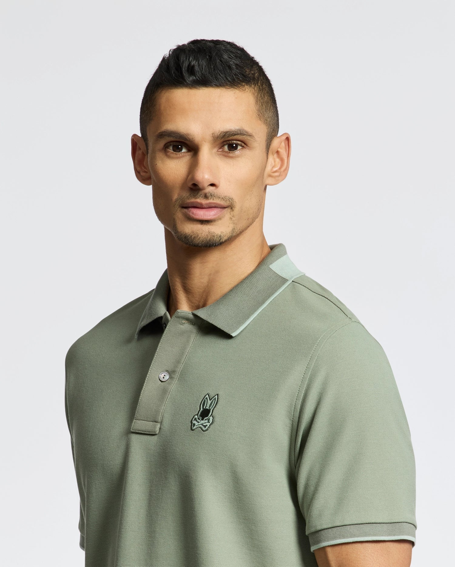 A person with short dark hair wears a muted green slim fit MENS NEW CASTLE PIQUE POLO by Psycho Bunny, crafted from Pima cotton with a small embroidered logo on the chest. The background is plain white.