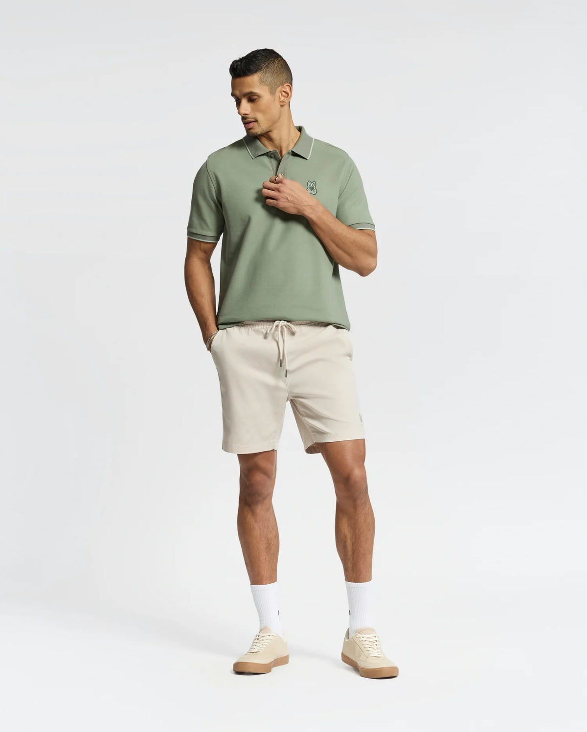 A man stands against a plain background wearing a light green Psycho Bunny Mens New Castle Pique Polo - B6K487F200 along with beige shorts, white socks, and beige sneakers. His hands are in his pockets as he gazes slightly downward.