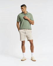 A man stands against a plain background wearing a light green Psycho Bunny Mens New Castle Pique Polo - B6K487F200 along with beige shorts, white socks, and beige sneakers. His hands are in his pockets as he gazes slightly downward.