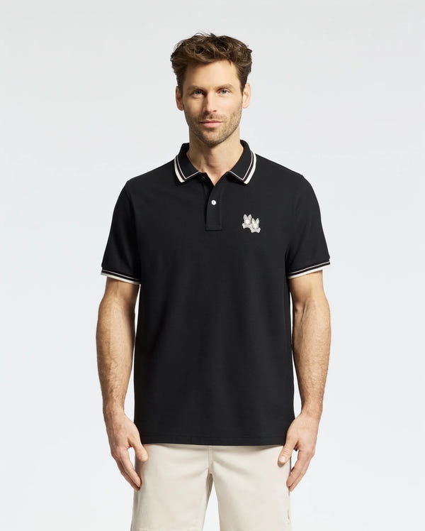 In a stylish Psycho Bunny Elliot Pique Polo (B6K505F200) crafted from luxurious Pima cotton, a man confidently stands against a plain white background. His polo features an embroidered Bunny on the chest and pairs perfectly with beige pants.