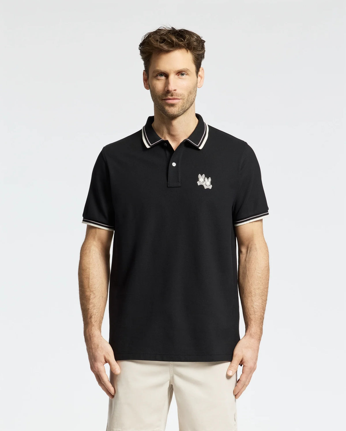 In a stylish Psycho Bunny Elliot Pique Polo (B6K505F200) crafted from luxurious Pima cotton, a man confidently stands against a plain white background. His polo features an embroidered Bunny on the chest and pairs perfectly with beige pants.