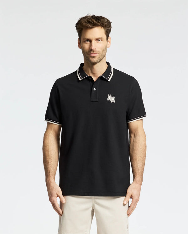 In a stylish Psycho Bunny Elliot Pique Polo (B6K505F200) crafted from luxurious Pima cotton, a man confidently stands against a plain white background. His polo features an embroidered Bunny on the chest and pairs perfectly with beige pants.