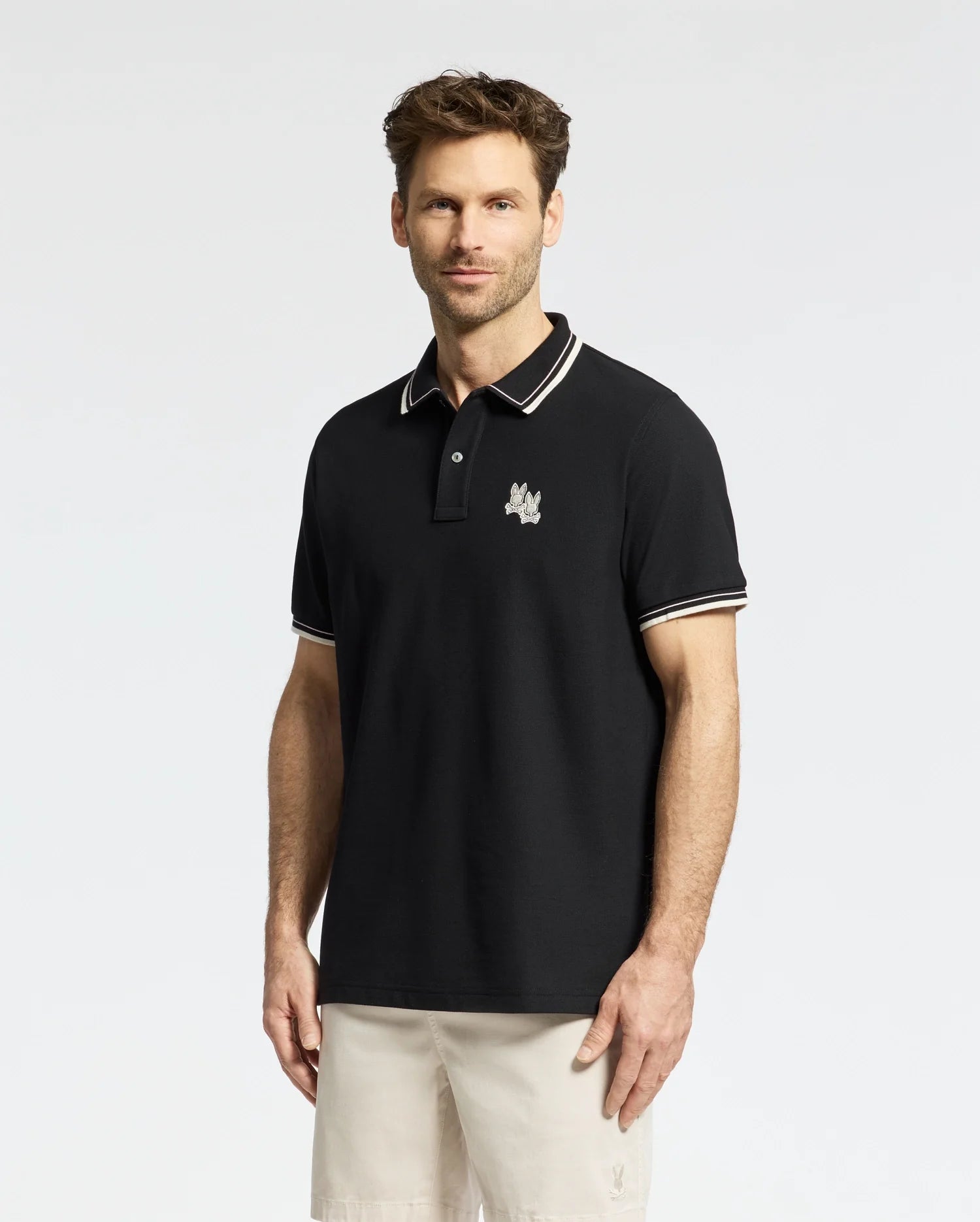A man sports the Psycho Bunny Men's Elliot Pique Polo (B6K505F200) in black Pima cotton with white trim on the collar and sleeves, highlighted by an embroidered Bunny logo on the left chest, paired with light shorts against a plain white backdrop.