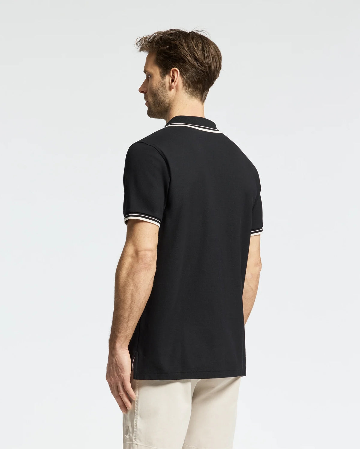 A person with short hair and fair skin is seen from behind, wearing a black MENS ELLIOT PIQUE POLO (B6K505F200) by Psycho Bunny, featuring white trim on the collar and sleeves. They are also sporting light-colored shorts against a plain, light background.