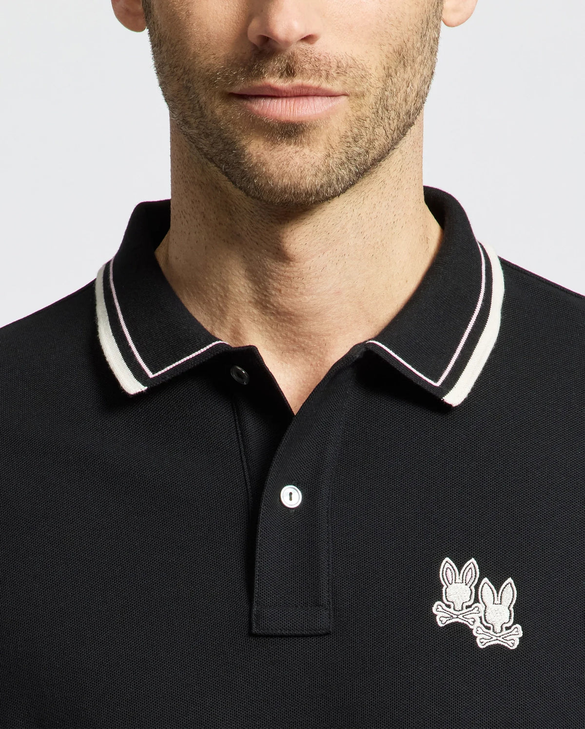A man dons the Psycho Bunny Men's Elliot Pique Polo (B6K505F200), crafted from soft Pima cotton. The black shirt features a white-striped collar and an embroidered Bunny logo of two bunnies with crossed bones near the collar, set against a plain, light-colored background.