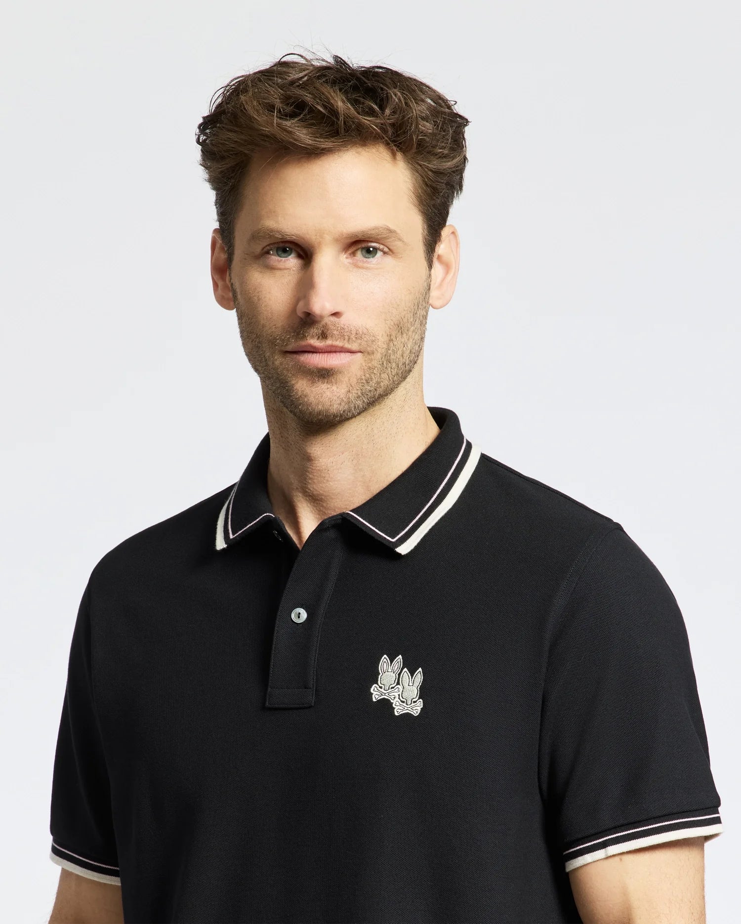 A man with short hair and facial hair is wearing the Psycho Bunny MENS ELLIOT PIQUE POLO (B6K505F200) in black, made from luxurious Pima cotton. It features an embroidered Bunny design near the collar and white trim on the collar and buttons, set against a plain white background.