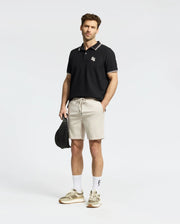A man with short hair stands against a plain background, wearing a Psycho Bunny men's Elliot Pique polo made of soft Pima cotton and beige shorts. He holds a black cap in his left hand, sporting white socks and beige sneakers.