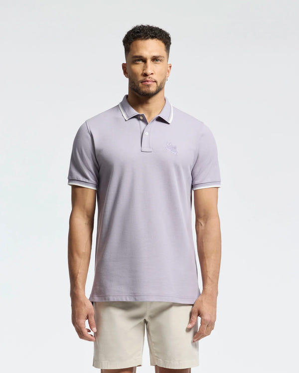 A man with a short beard wears a Psycho Bunny light gray MENS ELLIOT PIQUE POLO - B6K505F200 made of soft Pima cotton with a small chest embroidery, paired with beige shorts. He stands against a plain white background.