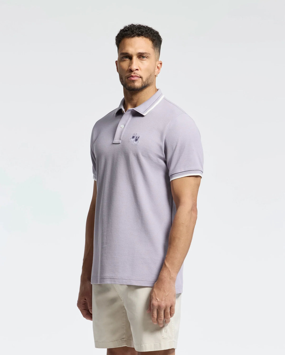 A man with short curly hair and a trimmed beard wears a light purple Psycho Bunny MENS ELLIOT PIQUE POLO - B6K505F200 in soft Pima cotton, featuring a subtle emblem. He pairs it with beige shorts against a plain white background.