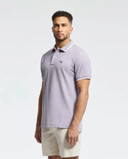 A man with short curly hair and a trimmed beard wears a light purple Psycho Bunny MENS ELLIOT PIQUE POLO - B6K505F200 in soft Pima cotton, featuring a subtle emblem. He pairs it with beige shorts against a plain white background.