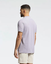 Man with short hair in a light purple Psycho Bunny Men's Elliot Pique Polo (B6K505F200), made from soft Pima cotton, paired with beige shorts, standing and facing left against a plain white background.