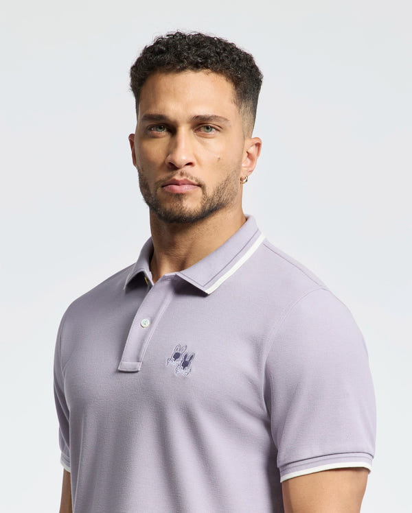 A person with short curly hair and a beard wears the Psycho Bunny MENS ELLIOT PIQUE POLO - B6K505F200, made from soft Pima cotton with an embroidered chest logo, standing against a plain background.