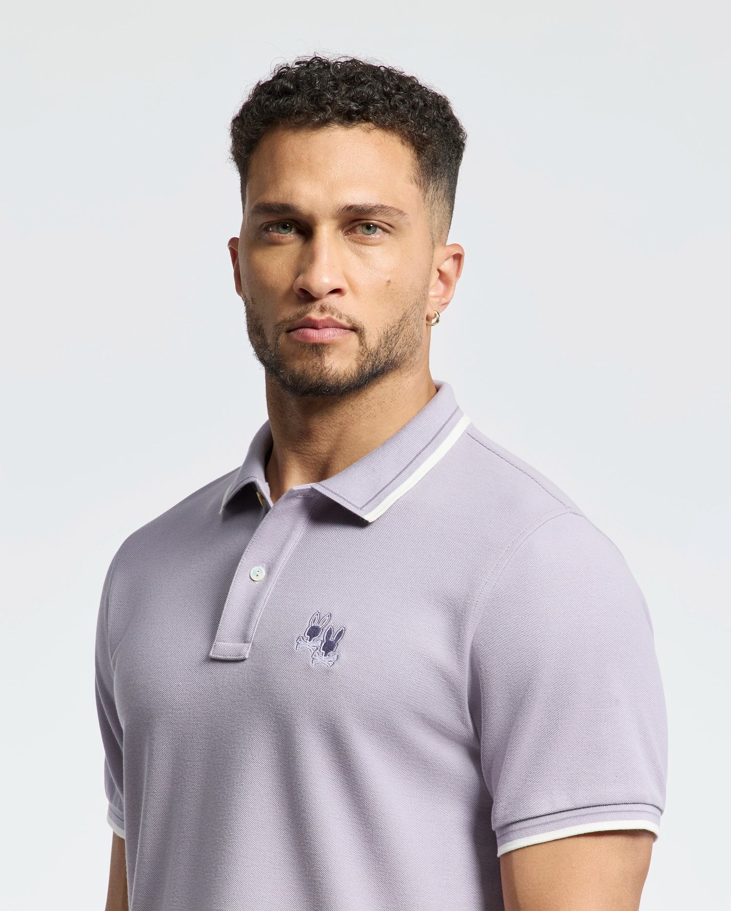 A person with short curly hair and a beard wears the Psycho Bunny MENS ELLIOT PIQUE POLO - B6K505F200, made from soft Pima cotton with an embroidered chest logo, standing against a plain background.