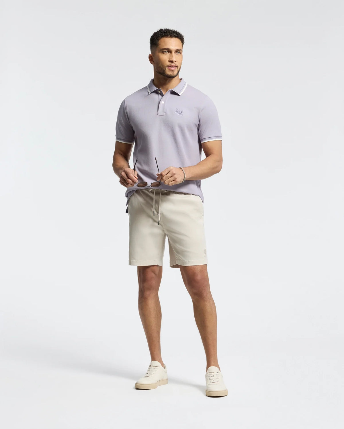 A man against a plain white background wears the light purple MENS ELLIOT PIQUE POLO by Psycho Bunny and beige shorts. He holds sunglasses and dons white sneakers, exuding a subtle yet bold style.
