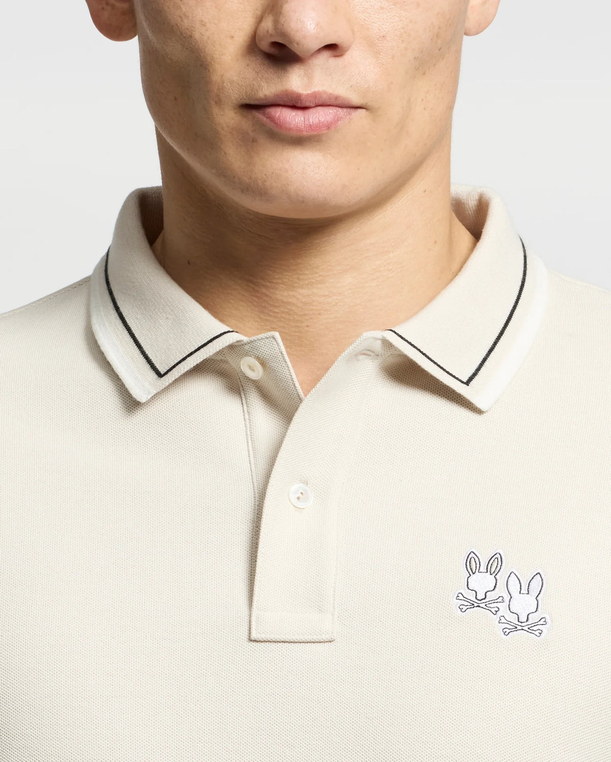 A person wearing a Psycho Bunny MENS ELLIOT PIQUE POLO - B6K505F200 in light beige, crafted from soft Pima cotton, featuring cartoon skulls and bunny ears on the left chest with a black-edged collar against a plain white background.