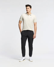 Against a plain backdrop, a man wears the Psycho Bunny MENS ELLIOT PIQUE POLO - B6K505F200 crafted from Pima cotton. He complements it with dark jogger pants and white sneakers, hands in pockets, looking sideways with effortless style.