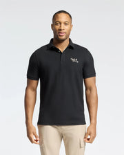 A man in a Psycho Bunny MENS DESERT PIQUE POLO (B6K506F200) with embroidered text on the chest stands against a light background. With short hair and beige pants, he exudes timeless style, hands at his sides, gazing directly at the camera.