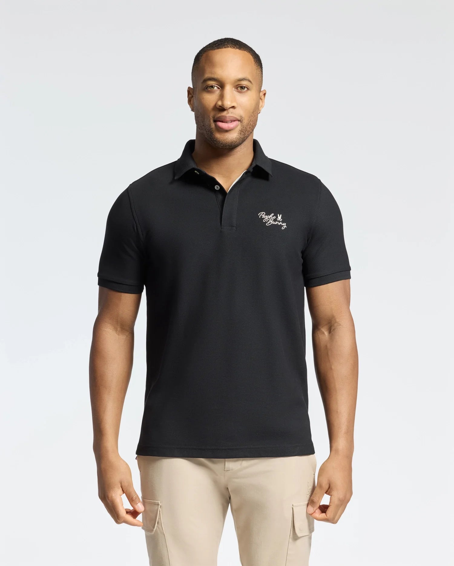 A man in a Psycho Bunny MENS DESERT PIQUE POLO (B6K506F200) with embroidered text on the chest stands against a light background. With short hair and beige pants, he exudes timeless style, hands at his sides, gazing directly at the camera.