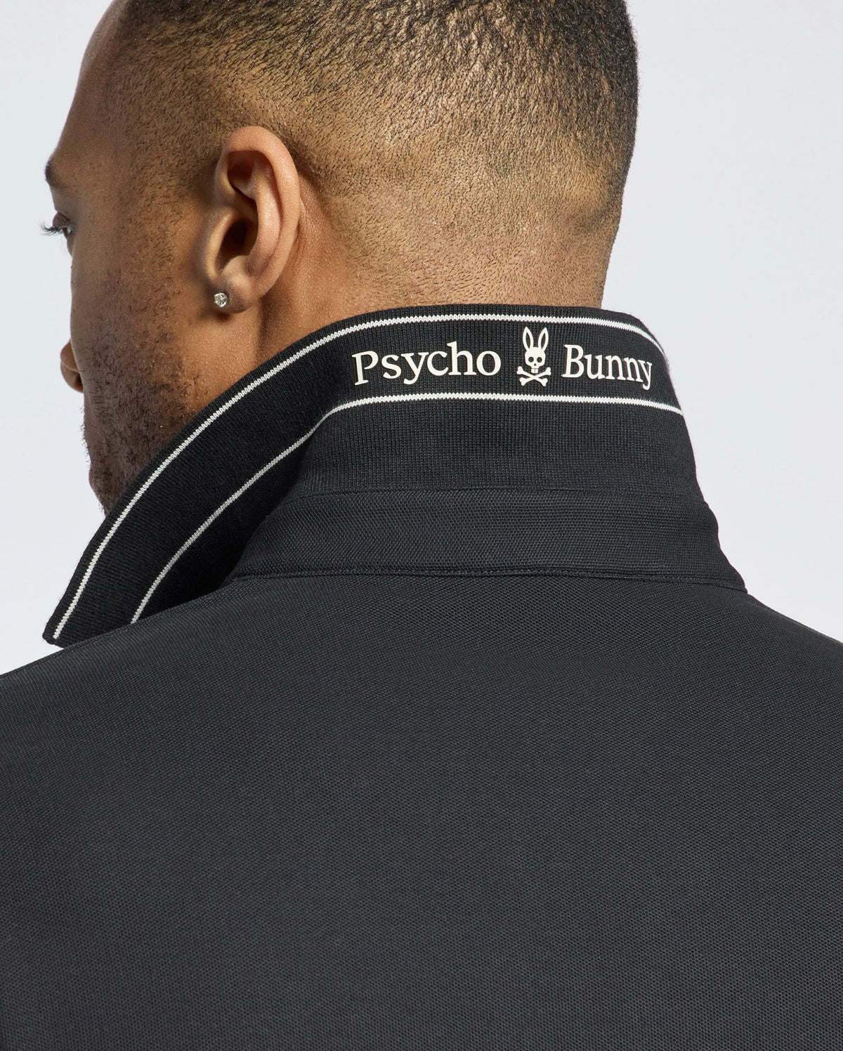 A person with short hair and a small earring wears the Psycho Bunny MENS DESERT PIQUE POLO - B6K506F200 in black, seen from the back. The turned-up collar reveals "Psycho Bunny" and a white bunny logo embroidered. Made of Pima cotton, this polo offers timeless style.