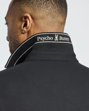 A person with short hair and a small earring wears the Psycho Bunny MENS DESERT PIQUE POLO - B6K506F200 in black, seen from the back. The turned-up collar reveals "Psycho Bunny" and a white bunny logo embroidered. Made of Pima cotton, this polo offers timeless style.