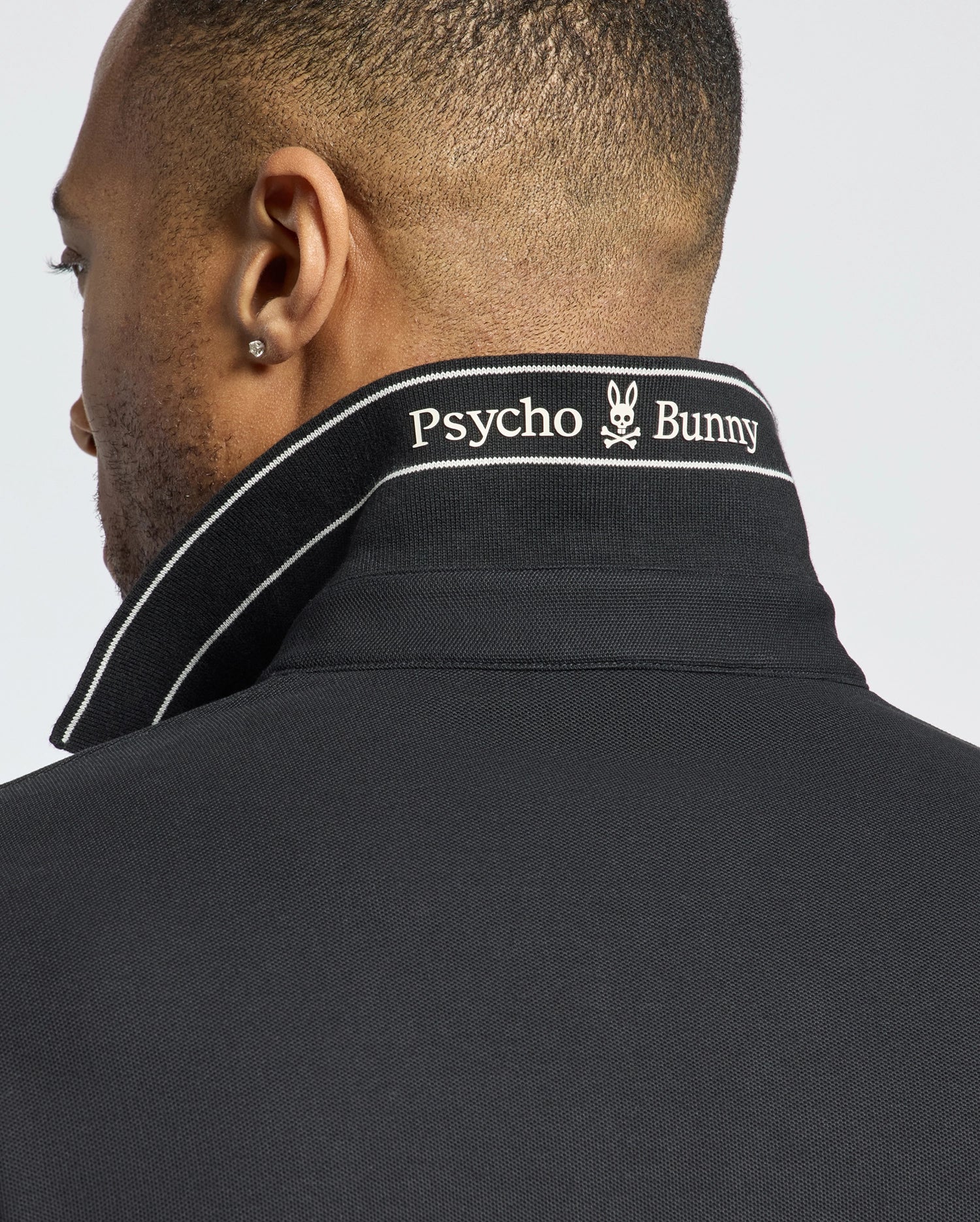 A person with short hair and a small earring wears the Psycho Bunny MENS DESERT PIQUE POLO - B6K506F200 in black, seen from the back. The turned-up collar reveals "Psycho Bunny" and a white bunny logo embroidered. Made of Pima cotton, this polo offers timeless style.
