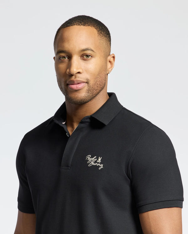A person wears a black Psycho Bunny MENS DESERT PIQUE POLO, made from luxurious Pima cotton with a small chest logo, against a neutral gray background as they confidently face the camera.