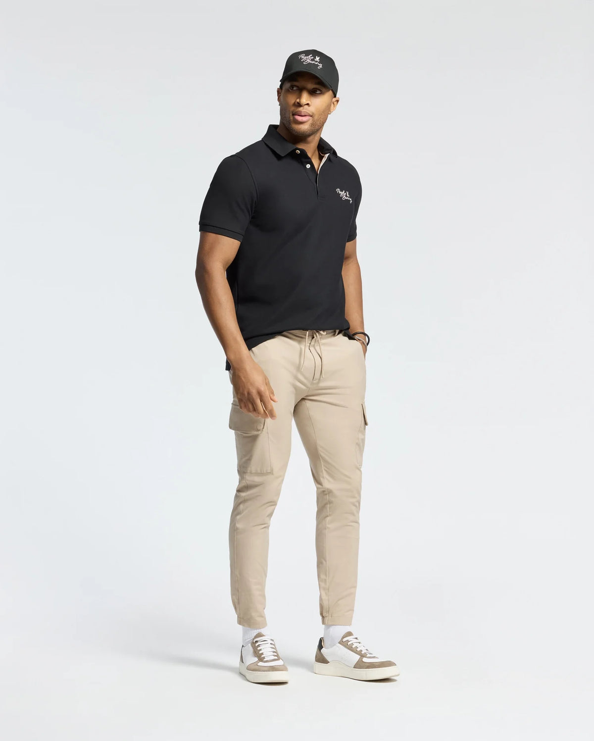 Someone in a Psycho Bunny MENS DESERT PIQUE POLO - B6K506F200 classic black, beige pants, and a matching black cap poses against a light background. They finish their timeless look with white sneakers featuring brown accents.