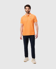 A man stands against a plain gray background, wearing an orange Psycho Bunny MENS BELTON PIQUE POLO - B6K575C200 with white trim and black pants. The embroidered Bunny logo adds a subtle touch of character. He completes the look with light-colored sneakers.