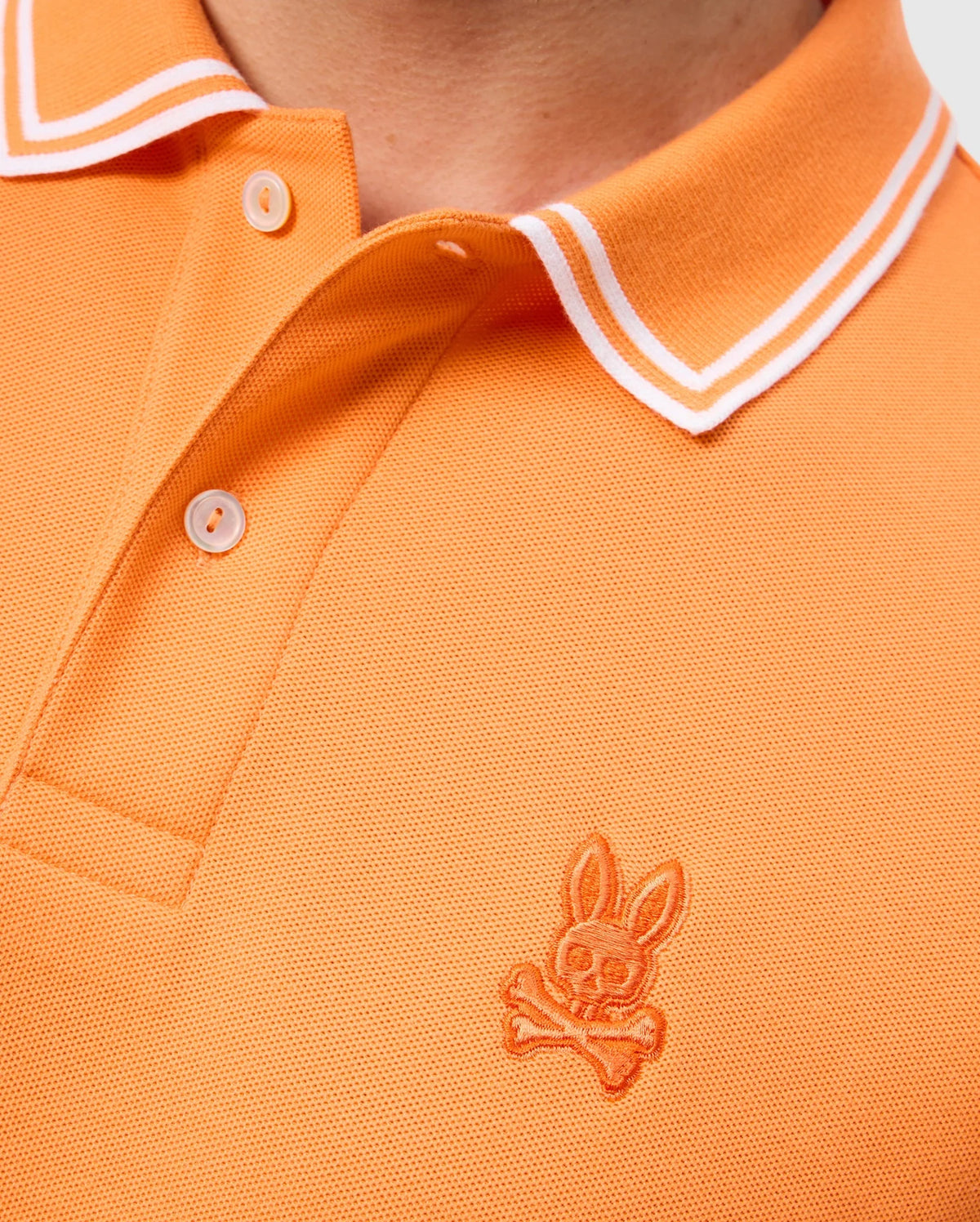Close-up of a person wearing an orange MENS BELTON PIQUE POLO - B6K575C200 by Psycho Bunny. The Pima cotton shirt features a two-button placket and white double stripes on the collar. An embroidered Bunny logo with a skull and crossbones is visible on the left chest.