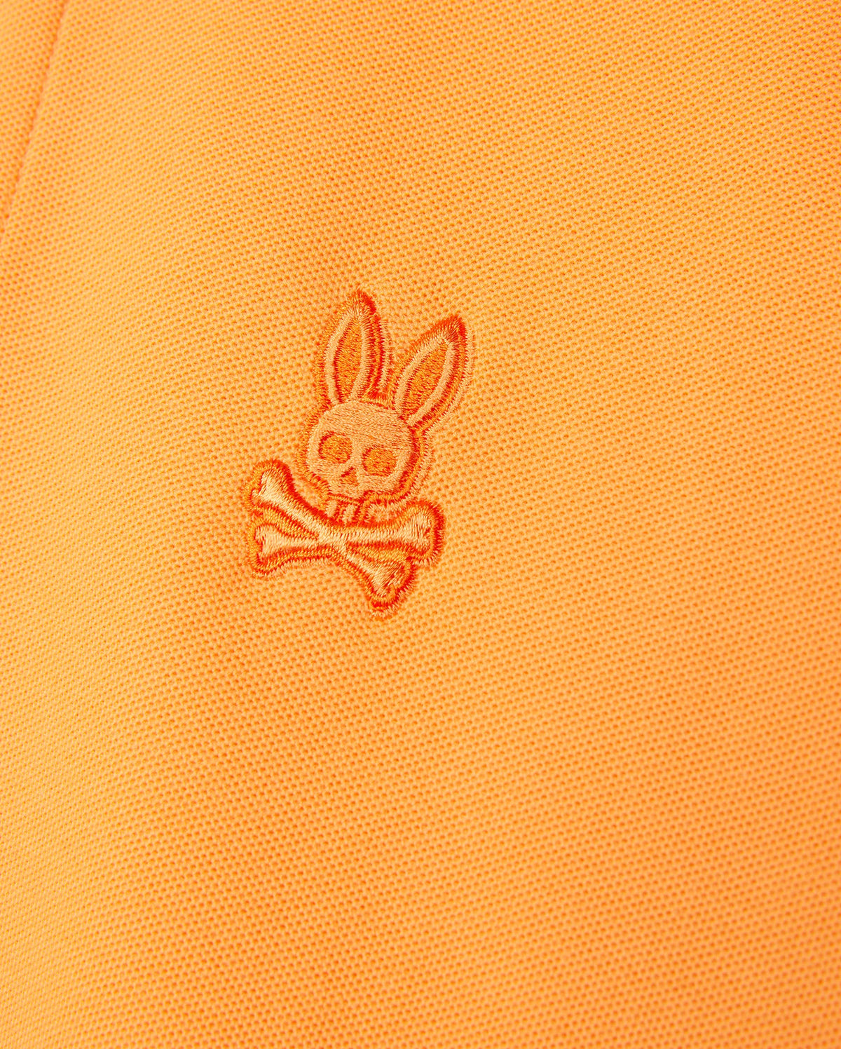 Close-up of an orange Psycho Bunny MENS BELTON PIQUE POLO - B6K575C200 made from Pima cotton featuring a stitched embroidered Bunny logo. The detailed embroidery showcases a stylized bunny skull with large ears above two crossed bones.