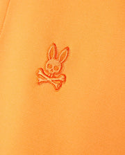 Close-up of an orange Psycho Bunny MENS BELTON PIQUE POLO - B6K575C200 made from Pima cotton featuring a stitched embroidered Bunny logo. The detailed embroidery showcases a stylized bunny skull with large ears above two crossed bones.