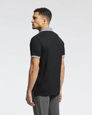 A man with short curly hair stands gazing sideways, wearing the Psycho Bunny MENS JONAS STRIPE COLLAR PIQUE POLO in black with grey trim, crafted from soft Pima cotton. His gray pants complement the look against a white background, subtly hinting at Peruvian elegance with their micro-stripe detail.