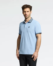 A man in a Psycho Bunny light blue MENS JONAS MICRO STRIPE COLLAR PIQUE POLO, marked with a black emblem on the left chest, and dark pants poses against a white background. The polo features micro-stripe detailing on its dark collar and sleeve trims. He has short brown hair and stubble.