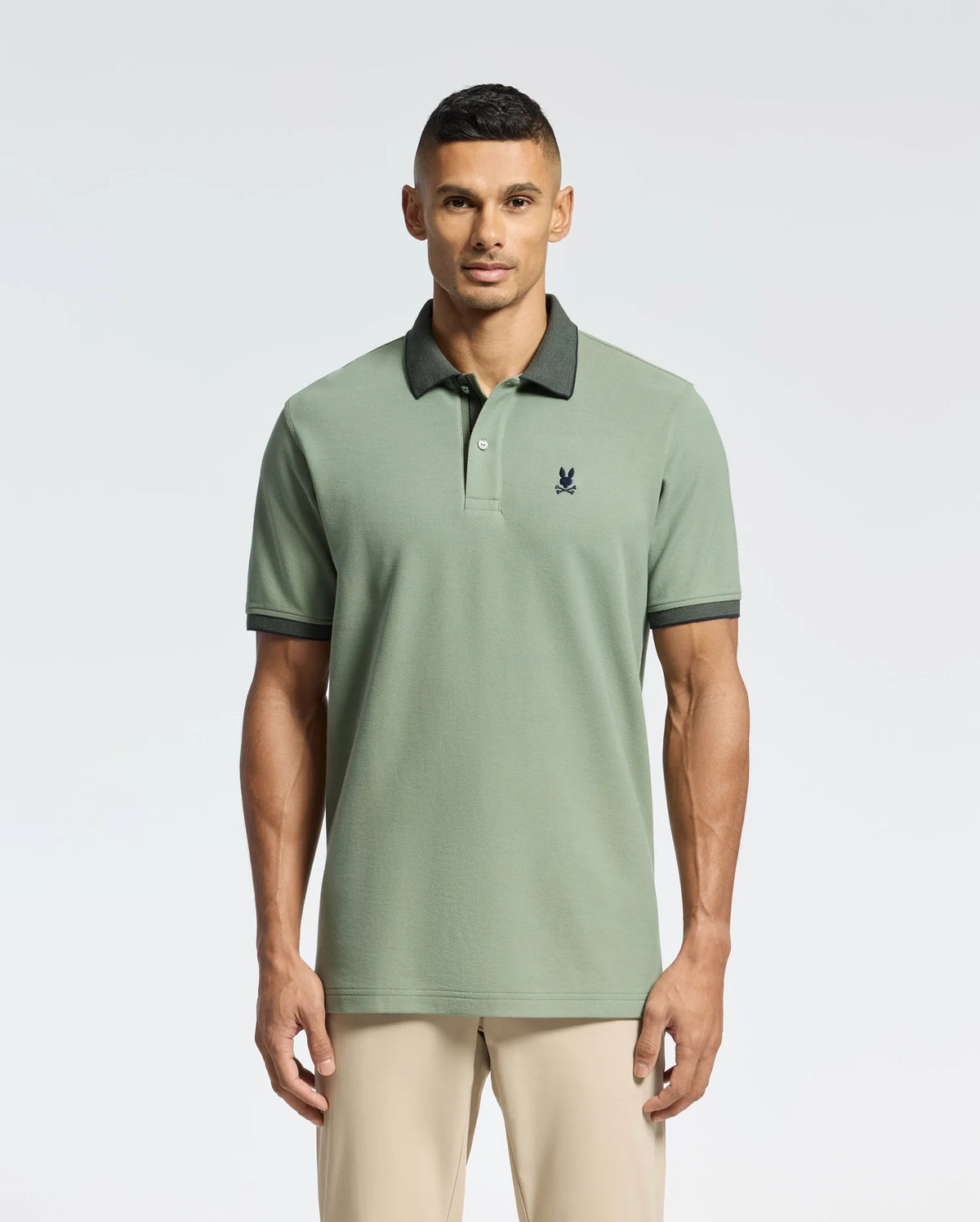 A man sports the Psycho Bunny MENS JONAS STRIPE COLLAR PIQUE POLO in sage green, made from soft Pima cotton. It features a dark collar and embroidered bunny emblem on the chest. He has short hair, poses against a plain background, and pairs it with light-colored pants.