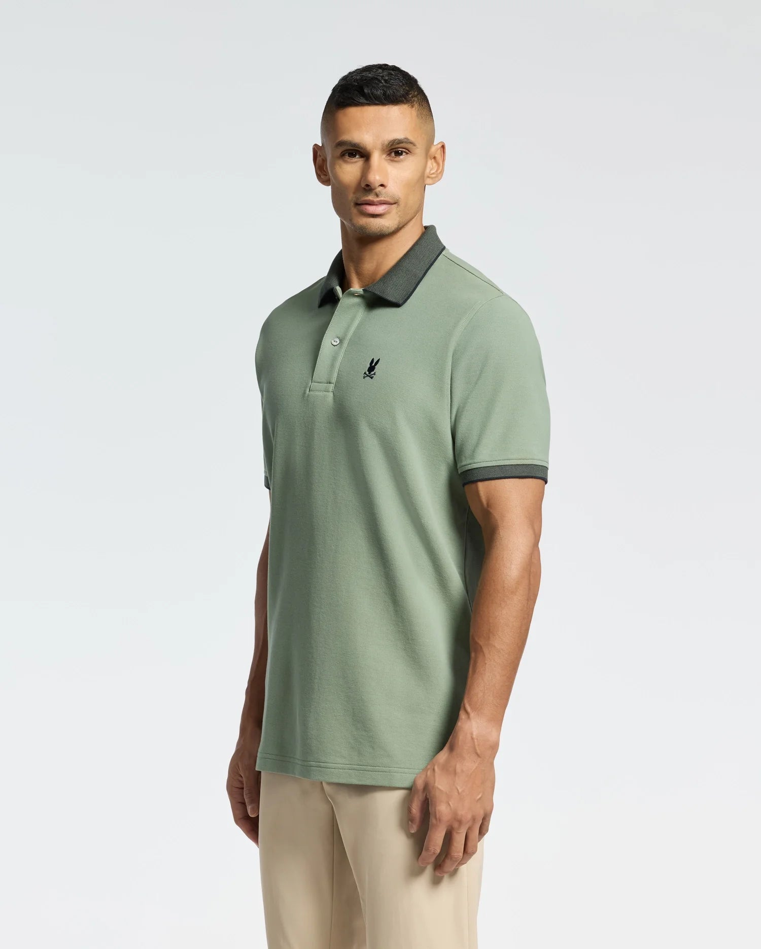 Wearing the MENS JONAS STRIPE COLLAR PIQUE POLO by Psycho Bunny in green, featuring a black collar and embroidered bunny logo, paired with beige pants, a person stands against a plain light background.